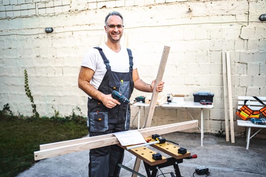 Mature male carpenter working with electrical screwdriver on a wooden plank outdoors.