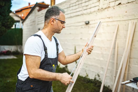 Mature male carpenter measuring wooden plank outdoors.