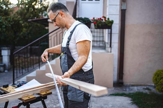 Mature male carpenter measuring wooden plank outdoors.