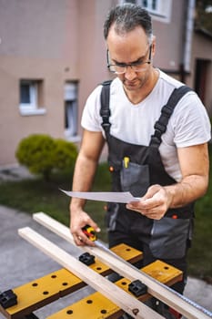 Mature male carpenter measuring wooden plank outdoors.