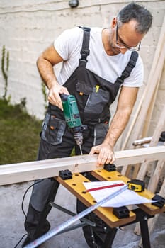 Mature male carpenter using electric screwdriver on a wooden plank outdoors.