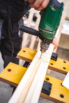 Unrecognizable male carpenter using electric drill on a wooden plank outdoors.