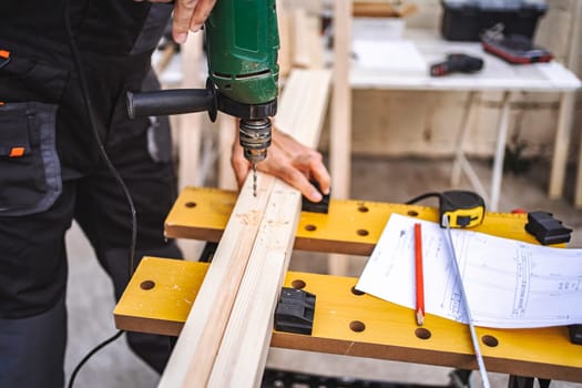 Unrecognizable male carpenter using electric drill on a wooden plank outdoors.