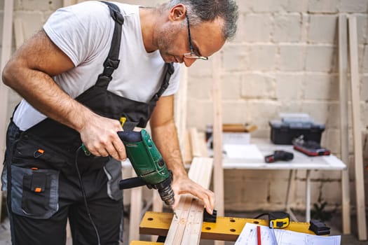 Mature male carpenter using electric screwdriver on a wooden plank outdoors.