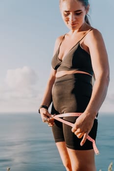 A woman in athletic wear measures her waist with a tape measure while standing on a cliff overlooking the ocean.