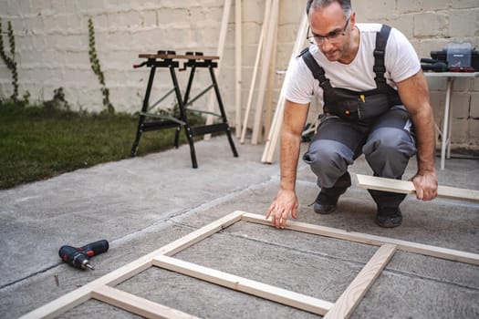 Mature male carpenter using electric screwdriver on a wooden plank outdoors.