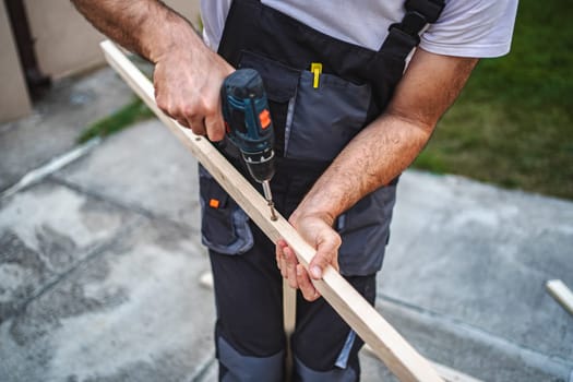 Unrecognizable male carpenter using electric screwdriver on a wooden plank outdoors.