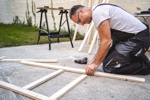Mature male carpenter using electric screwdriver on a wooden plank outdoors.