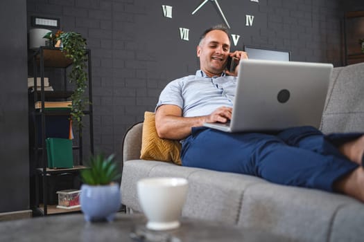 Middle aged Caucasian man talking on the phone while using laptop on the sofa.
