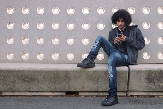 african american teenager with curly hair listening to music on his mobile phone, concept of technology of communication and urban lifestyle