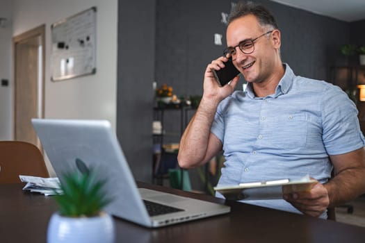 Middle aged Caucasian man talking on the phone while working on laptop at home.