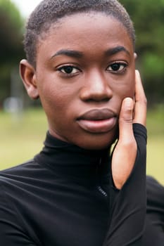 young tender ethnic female in black apparel with short hair looking at camera touching face with hand