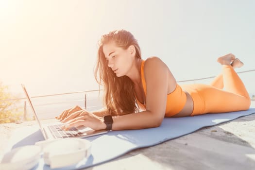 A woman in orange sportswear lies on a yoga mat outdoors, using a laptop.