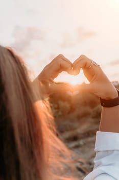 A woman forms a heart shape with her hands in front of a setting sun.