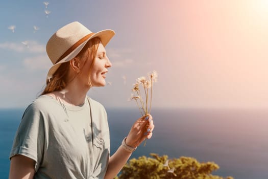 A woman with red hair blows on dandelions while wearing a straw hat, her back is to the sun