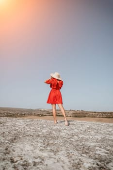 A young woman stands on a rocky outcrop wearing a red dress and a straw hat, looking out at the horizon.