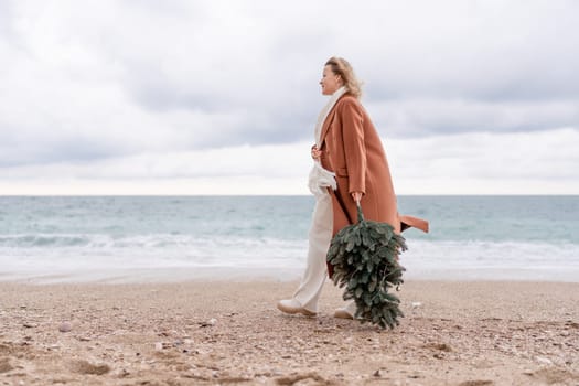 Blond woman Christmas sea. Christmas portrait of a happy woman walking along the beach and holding a Christmas tree in her hands. She is wearing a brown coat and a white suit