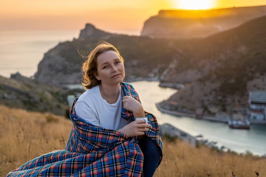 A woman is sitting on a hill with a blanket wrapped around her. She is looking out at the ocean