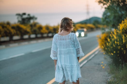 A woman in a white dress is standing on the side of a road. The road is empty and there is a car in the background. The woman is looking off into the distance, possibly lost in thought