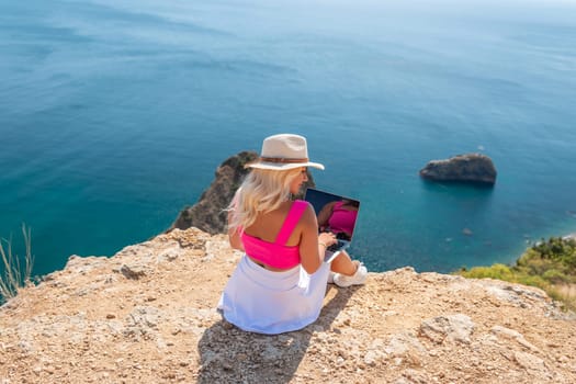 A woman is sitting on a rocky beach with a laptop in front of her. She is wearing a pink top and a straw hat. The beach is near the ocean, and the sky is clear