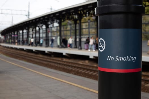 No smoking sign at stop of passenger trains, inscription on pole prohibiting smoking on the platform against the backdrop of stop for train with large number of passengers
