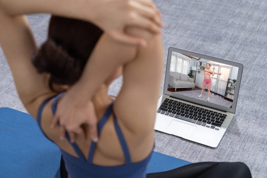 Asian woman in sportswear doing yoga exercise on fitness mat as her home workout training routine. Healthy body care lifestyle woman watching online yoga video on laptop. Vigorous