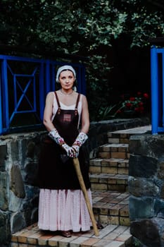 Woman dressed in a long pink dress and leather apron stands confidently against a stone wall holding axe