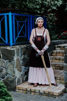 Woman dressed in a long pink dress and leather apron stands confidently against a stone wall holding axe