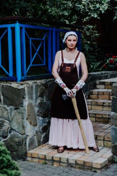 Woman dressed in a long pink dress and leather apron stands confidently against a stone wall holding axe