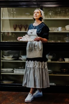 A woman in a vintage apron holds a silver tray filled with beautifully arranged marshmallow in a cozy kitchen. She stands in front of a glass cabinet filled with elegant glassware, looking off to the side with a calm, content expression.