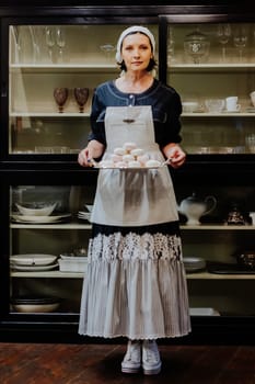 A woman in a vintage apron holds a silver tray filled with beautifully arranged marshmallow in a cozy kitchen. She stands in front of a glass cabinet filled with elegant glassware, looking off to the side with a calm, content expression.