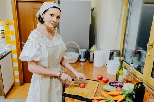A woman is preparing a meal in a cozy kitchen. She is chopping fresh tomatoes on a wooden cutting board, surrounded by an array of colorful vegetables such as carrots, cauliflower, and cucumbers.