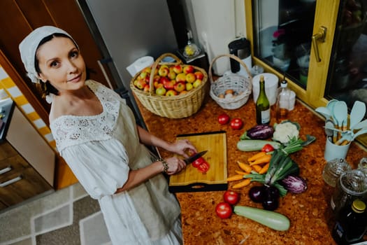 A woman is preparing a meal in a cozy kitchen. She is chopping fresh tomatoes on a wooden cutting board, surrounded by an array of colorful vegetables such as carrots, cauliflower, and cucumbers.