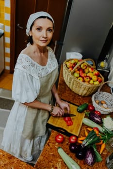 A woman is preparing a meal in a cozy kitchen. She is chopping fresh tomatoes on a wooden cutting board, surrounded by an array of colorful vegetables such as carrots, cauliflower, and cucumbers.