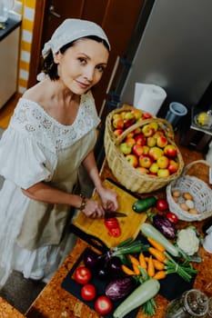 A woman is preparing a meal in a cozy kitchen. She is chopping fresh tomatoes on a wooden cutting board, surrounded by an array of colorful vegetables such as carrots, cauliflower, and cucumbers.