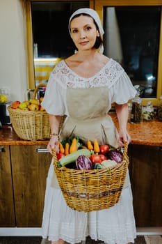 Woman dressed in a traditional white dress and apron holds a large wicker basket filled with fresh vegetables, including carrots, tomatoes, and eggplants, in a cozy kitchen.