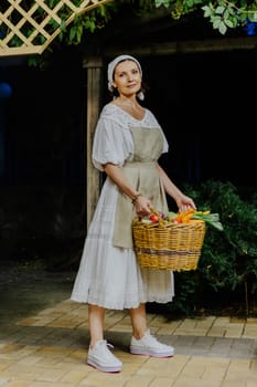 Woman dressed in a traditional white dress and apron holds a large wicker basket filled with fresh vegetables, including carrots, tomatoes, and eggplants, in a cozy kitchen.