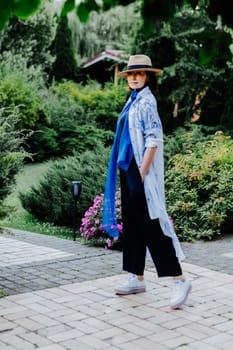 Fashionable woman is seen walking outdoors, dressed in a stylish combination of a long, flowing patterned coat, a blue scarf, wide-leg black pants, white sneakers, and a straw hat