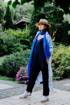 Fashionable woman is seen walking outdoors, dressed in a stylish combination of a long, flowing patterned coat, a blue scarf, wide-leg black pants, white sneakers, and a straw hat
