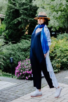 Fashionable woman is seen walking outdoors, dressed in a stylish combination of a long, flowing patterned coat, a blue scarf, wide-leg black pants, white sneakers, and a straw hat
