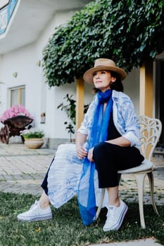 Fashionable woman is seen walking outdoors, dressed in a stylish combination of a long, flowing patterned coat, a blue scarf, wide-leg black pants, white sneakers, and a straw hat