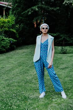 A fashionable woman stands confidently on a green lawn, wearing a bold, geometric-patterned blue jumpsuit, a light sleeveless vest, and white sneakers.