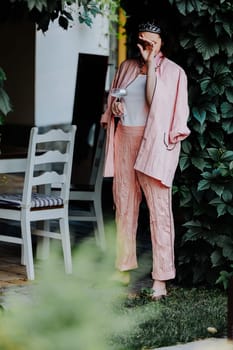 Fashionable woman dressed in a stylish light pink ensemble, including a satin jacket and matching pants, is standing outdoors under a canopy of leaves, holding a glass