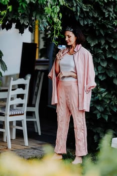 Fashionable woman dressed in a stylish light pink ensemble, including a satin jacket and matching pants, is standing outdoors under a canopy of leaves, holding a glass