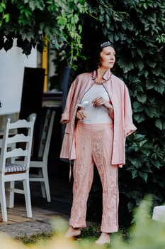 Fashionable woman dressed in a stylish light pink ensemble, including a satin jacket and matching pants, is standing outdoors under a canopy of leaves, holding a glass