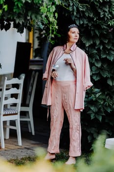 Fashionable woman dressed in a stylish light pink ensemble, including a satin jacket and matching pants, is standing outdoors under a canopy of leaves, holding a glass
