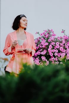 Serene woman stands outdoors next to a vibrant flower bush, dressed in a silky pink robe and holding a teacup