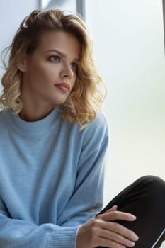 Beautiful blonde wearing nice blue sweater sitting on window sill looking out the window romantic studio portrait
