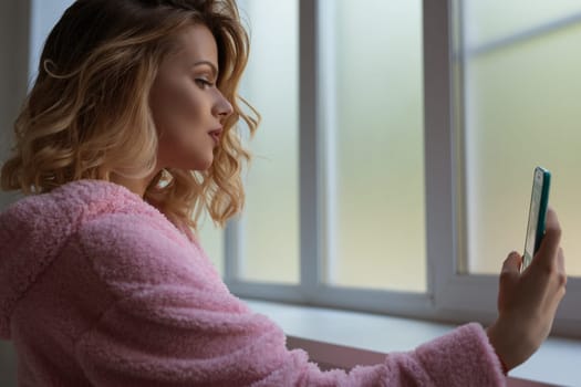 Gorgeous curly blonde in pink bathrobe making selfie near window sill