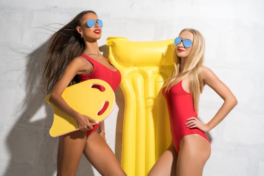 Two sexy girls wearing sunglasses, red swimsuit and summer hat posing in studio near brick white wall holding swimming air mattress